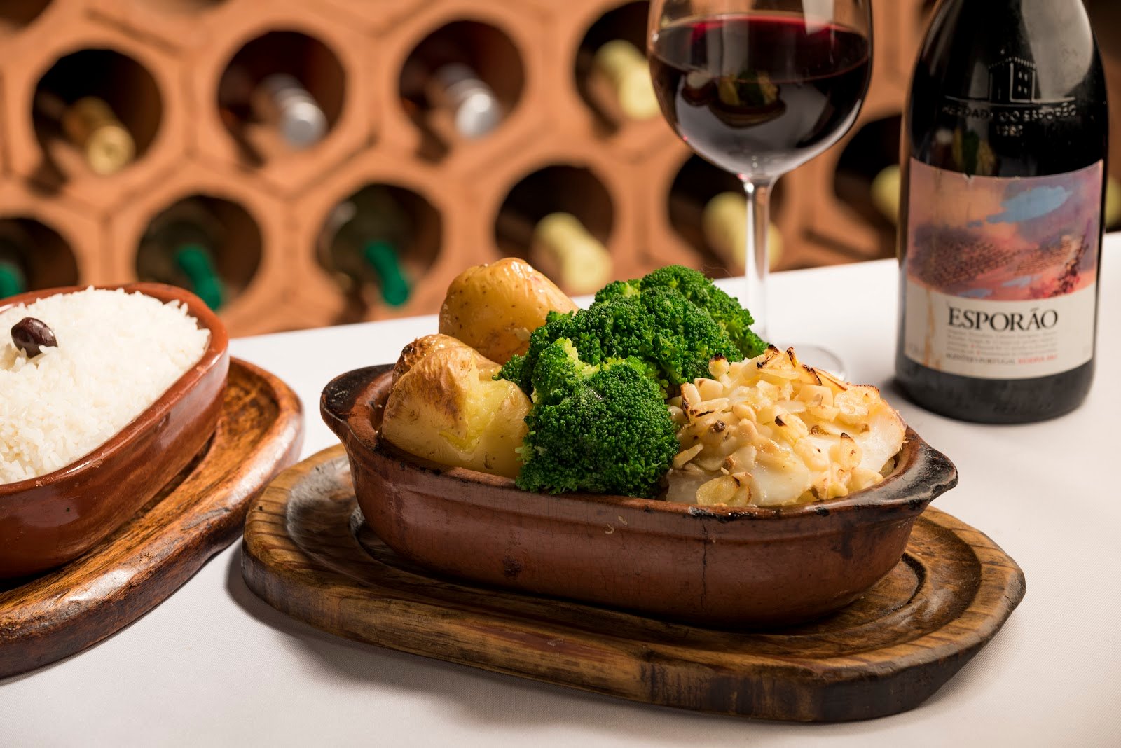 Bacalhau Ora Pois em travessa de barro, com brócolis, batatas ao murro, arroz e um copo de vinho tinto